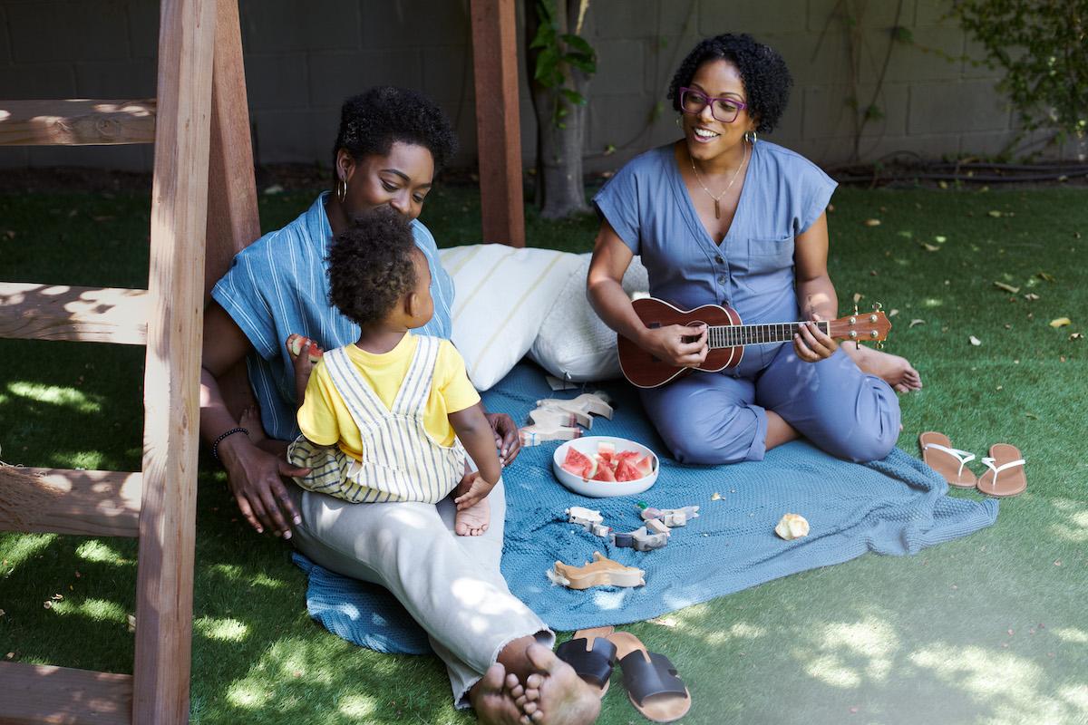 Two women and a child sit on a picnic blanket outdoors playing a ukulele