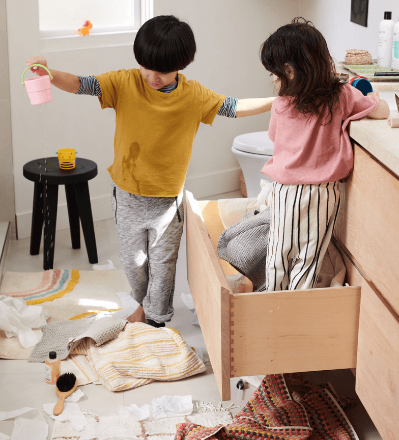 Two children stand over a mess of towels, toilet paper, and toys in a bathroom