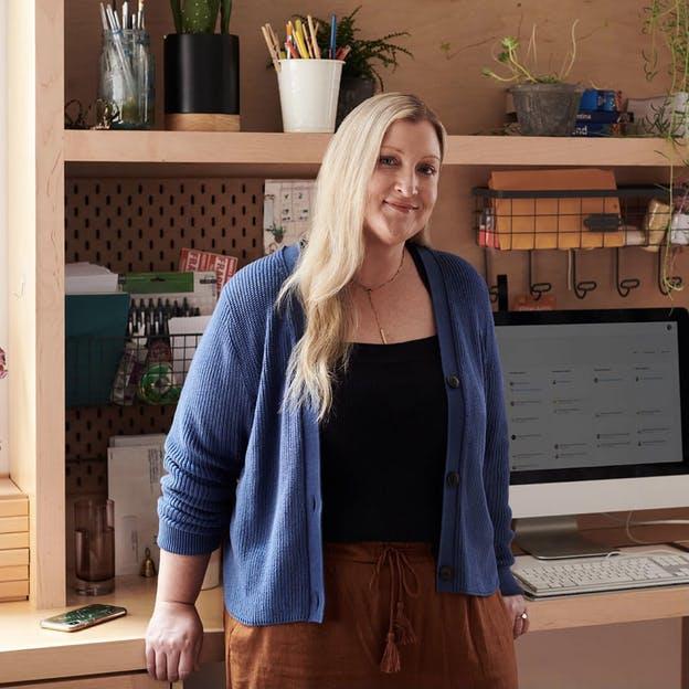 Personal assistant leans against home office workstation with shelves, a computer, plants, and envelopes