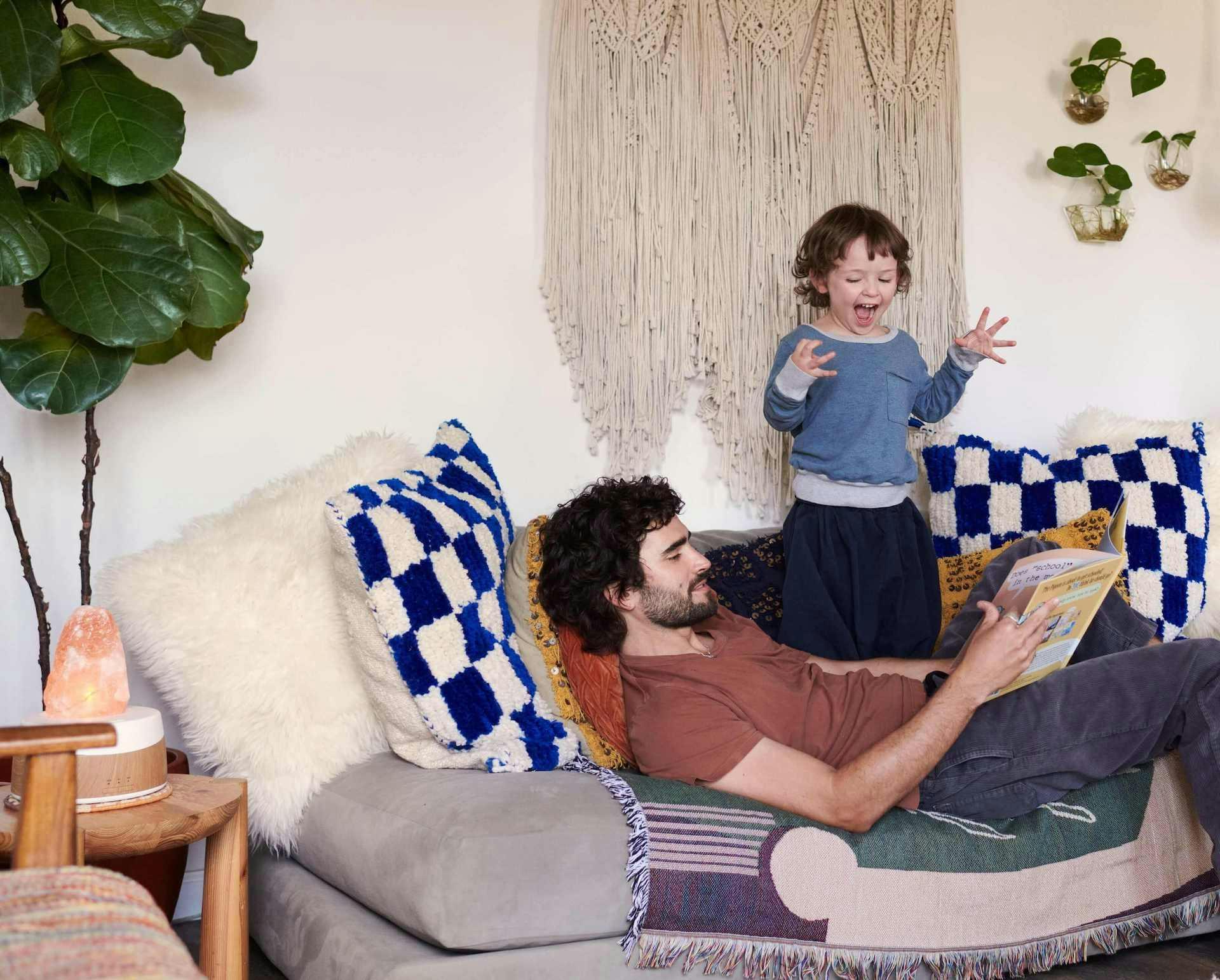 Man reclines on a couch while reading to his son who gestures in excitement